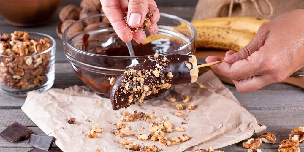 Økologisk banan dyppet i chokolade og drysset med karameliserede nødder. I baggrunden ser vi bananer, valnødder og chokolade, mens en person holder bananen, som er sat på en pind.