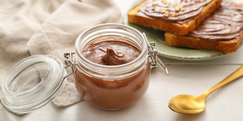 Økologisk chokoladesmør med hasselnødder og en gylden ske ved siden af på et bord med en beige dug i baggrunden og en tallerken med to brødskiver med chokoladesmørret.