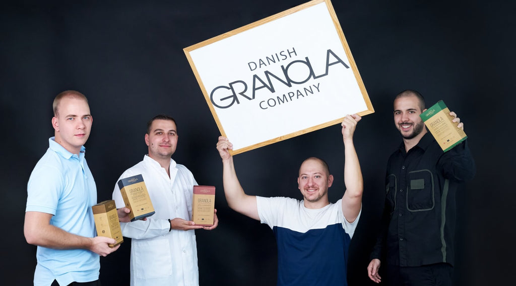 Holdet bag Danish Granola Company. Der er fire mænd på billedet - tre af dem holder granola kasser, og den fjerde holder et hvidt bræt med Danish Granola Company-logoet på det.