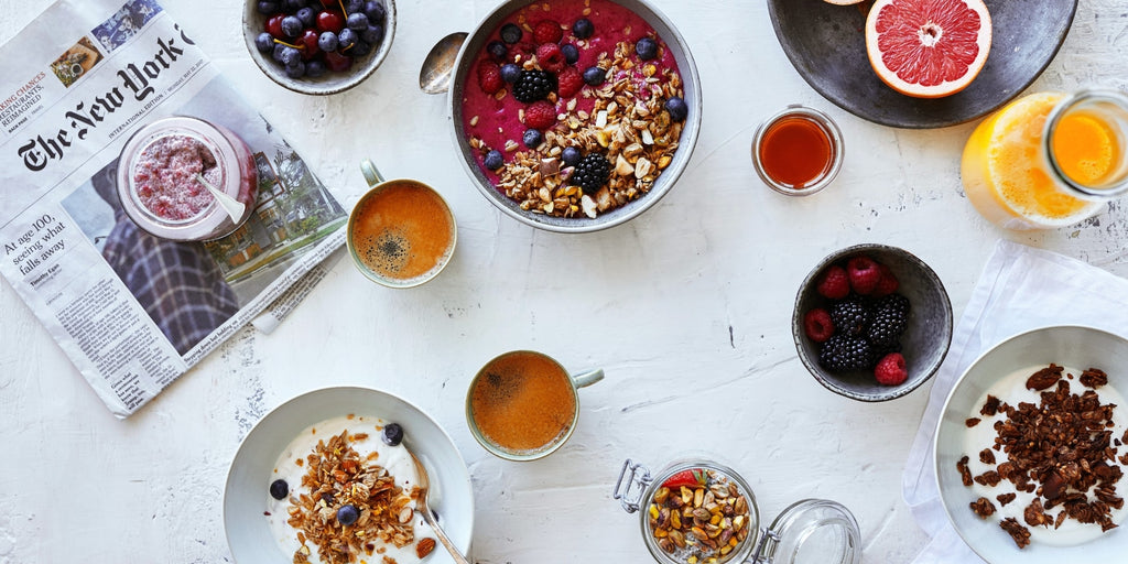 Blanding af produkter og smoothie-skåle med granola og bær. Der er også en New York Times avis, kopper med kaffe, appelsinjuice og noget frugt.