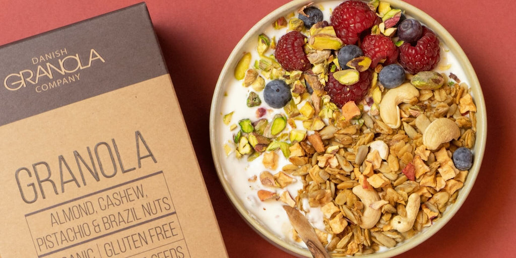 Økologisk granola med mandler, cashewnødder, pistacienødder, paranødder fra Danish Granola Company, med en smoothie-skål ved siden af på en rød baggrund.
