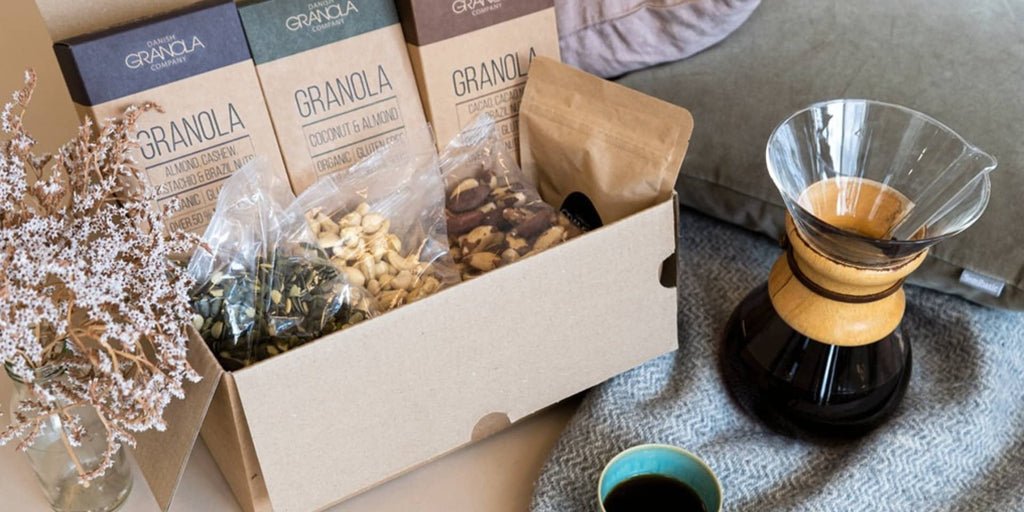 Morgen-Box fra Danish Granola Company med tre forskellige smagsvarianter i en æske sammen med nogle nødder og frø. Ved siden af æsken ser vi blomster og en kaffekande samt en kop kaffe.