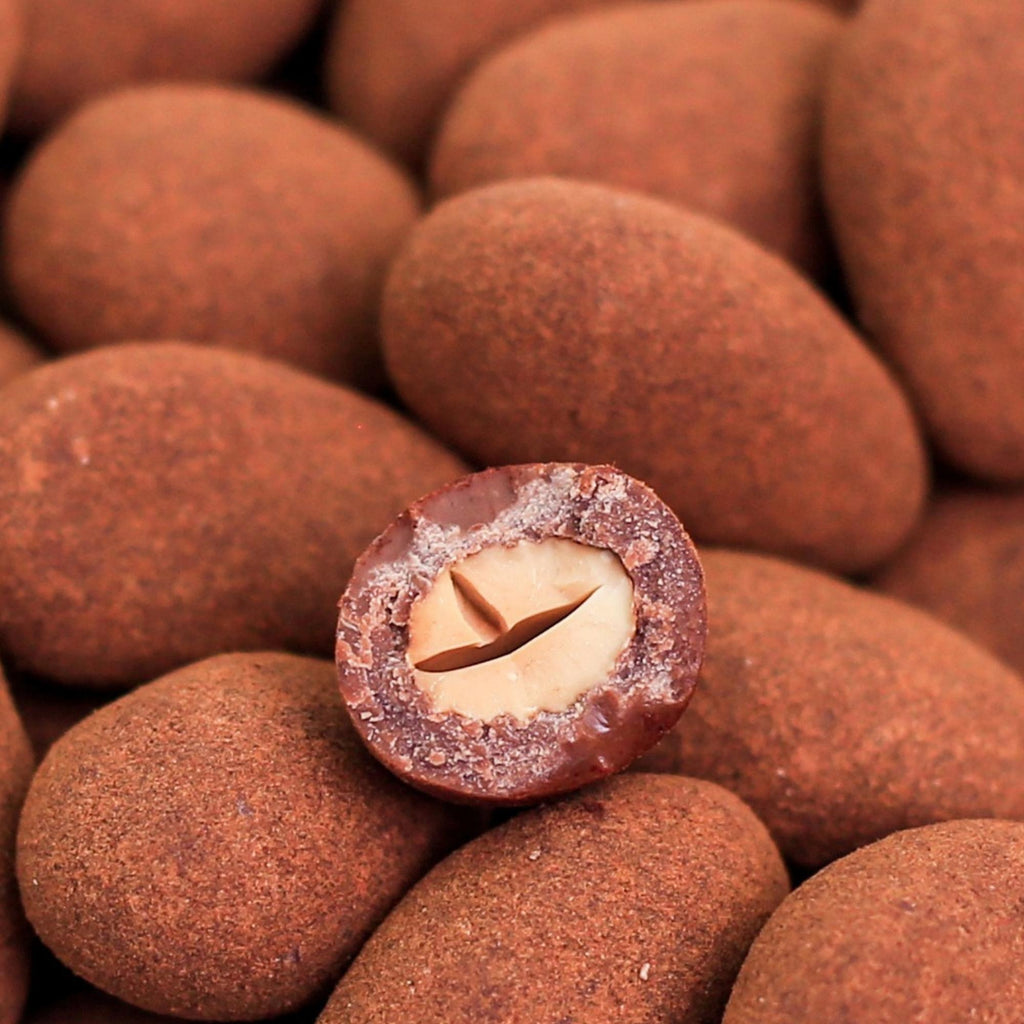 ØKO Chokoladeovertrukket mandler med kanel, billedet er med hvid baggrund, hvor der er zoomet ind på mandlerne. Dertil er der en mandel, som er skåret over så man kan se chokoladen omkring mandlen. 